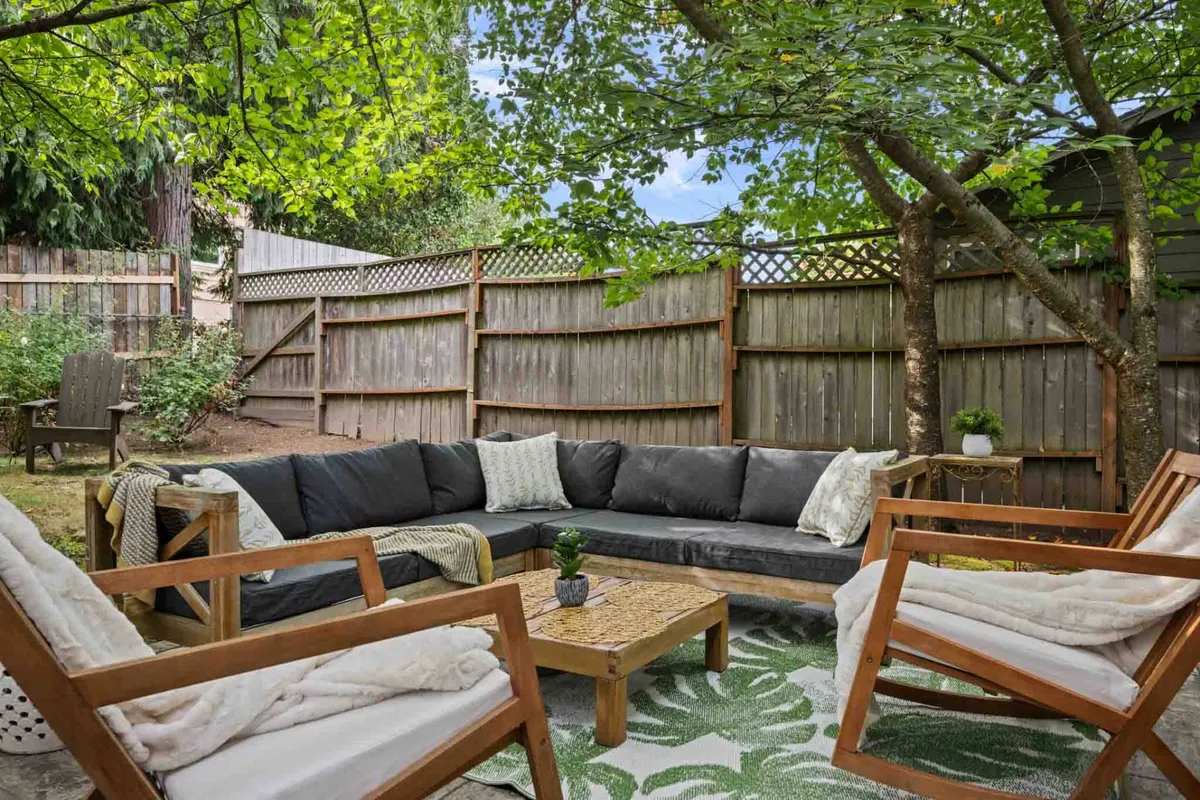 An outdoor patio setup featuring a sectional sofa, wooden chairs, a coffee table, and abundant greenery. The arrangement is cozy and inviting, ideal for relaxing or unwinding under the stars. The surrounding plants add to the sense of seclusion and privacy, with enough space for couples to enjoy a peaceful evening or morning coffee outdoors.