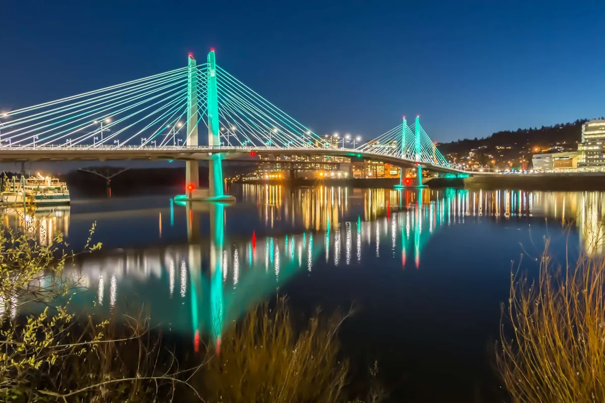 An illuminated bridge over the river with the reflections of city lights sparkling in the water. The image captures Portland's lively nighttime atmosphere and urban charm.