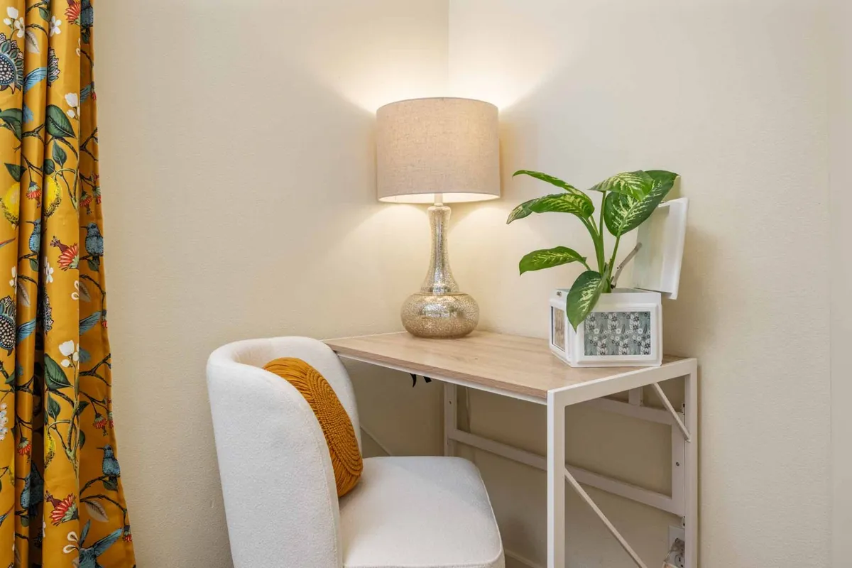 A calming interior view featuring a desk positioned by a window, flanked by gentle curtains and a potted plant. A soft, inviting lamp provides additional lighting, setting up a tranquil atmosphere ideal for work, study, or reading. The desk is uncluttered, promoting focus and productivity.