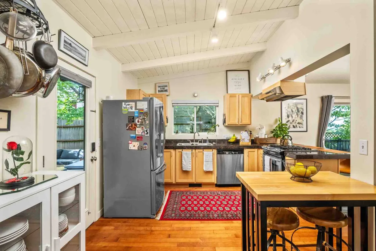 A warm, inviting space with a kitchen island in the foreground, a dining area just beyond, and large windows bringing in natural light and views of greenery. The fridge with magnets adds a personal, lived-in touch, emphasizing comfort and family life.