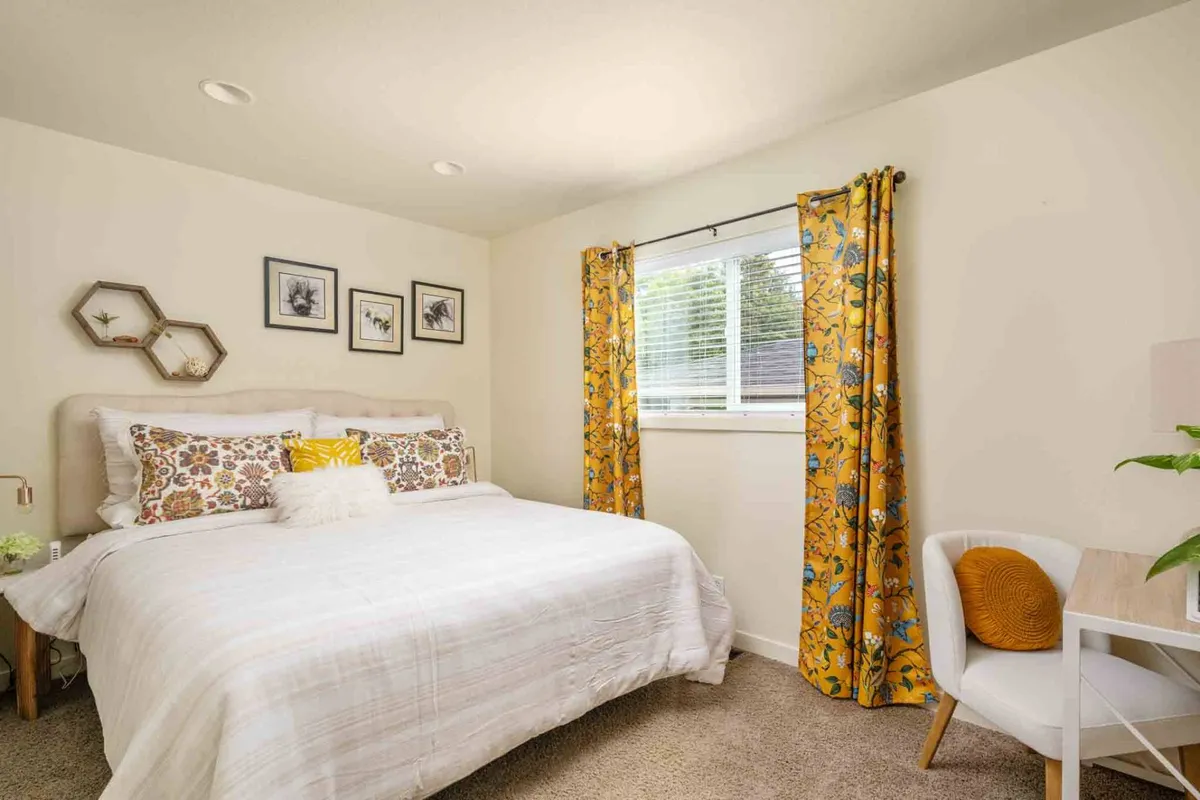 A cozy bedroom featuring a neatly made bed adorned with decorative pillows, colorful curtains that add cheerful vibrancy to the space, and framed wall art. The room is softly lit, creating an inviting ambiance perfect for relaxing or reading.
