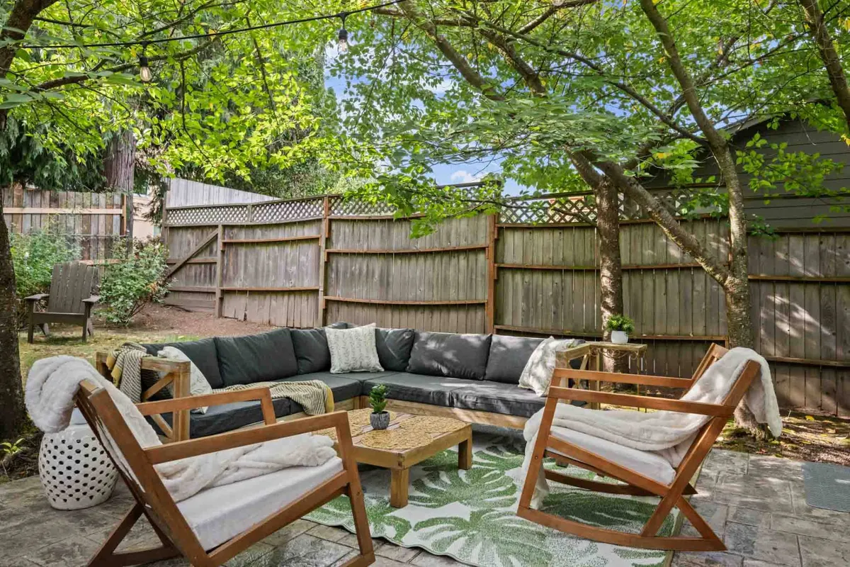 An inviting backyard patio set up with an outdoor seating area, framed by vibrant green foliage and a wooden fence. Decorative elements like a rug and cozy seating arrangements create a welcoming space for socializing or quiet reflection in the fresh air.
