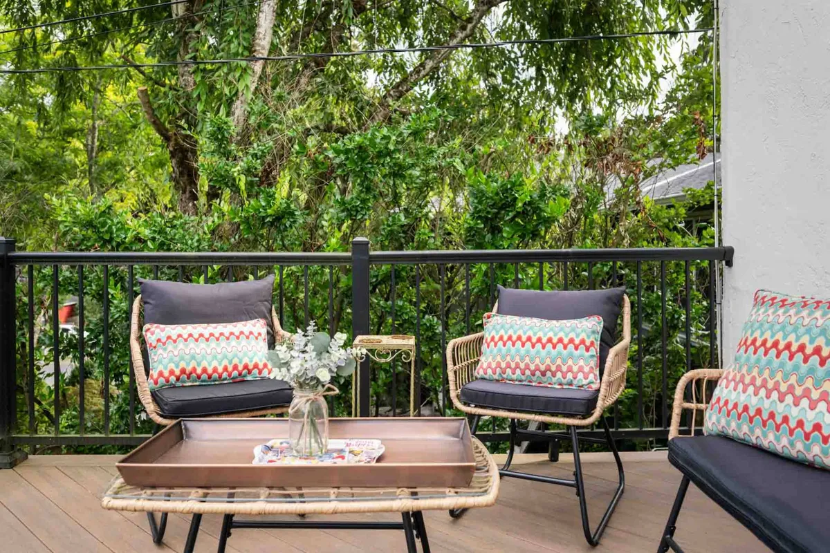An inviting outdoor setting featuring rattan chairs with decorative cushions and a coffee table with a vase, backed by lush greenery. The arrangement suggests a serene spot for morning coffee rituals, reading, or unwinding after a productive workday.