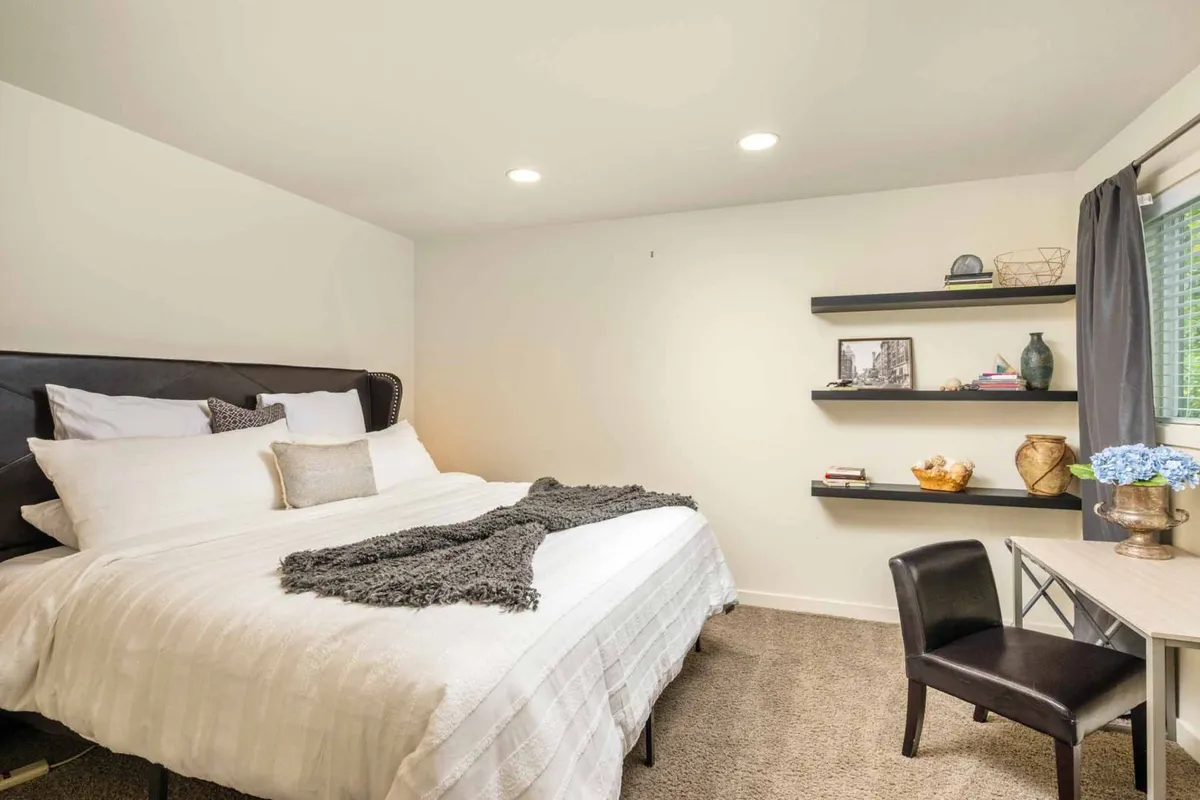 A tranquil bedroom equipped with a bed, neatly arranged shelves, a desk, and a chair. The workspace is integrated into the room, allowing for both relaxation and productivity, with soft lighting enhancing the serene mood.