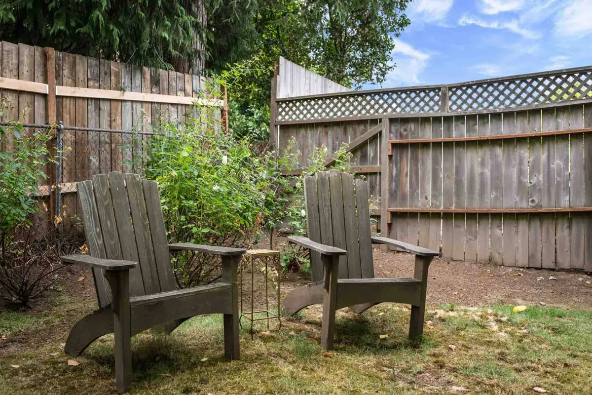 A quiet outdoor scene with two wooden Adirondack chairs placed amidst lush green foliage, enclosed by a wooden fence. The setup suggests a private retreat ideal for quiet conversation, reading, or simply enjoying nature in a peaceful backyard setting.