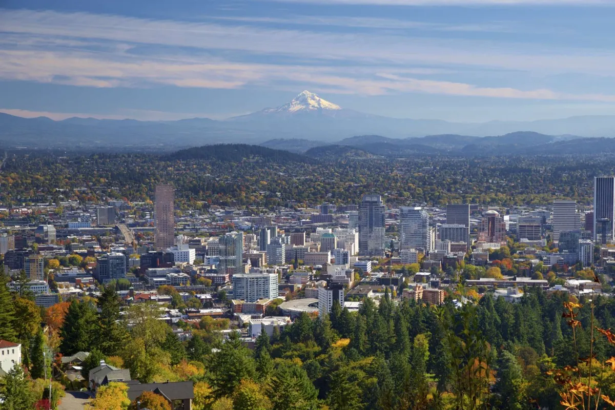 A sweeping view of downtown Portland with Mount Hood in the background and tree-covered hills surrounding the city. The image captures the city's skyline at sunrise, with fall foliage adding a burst of color to the landscape.