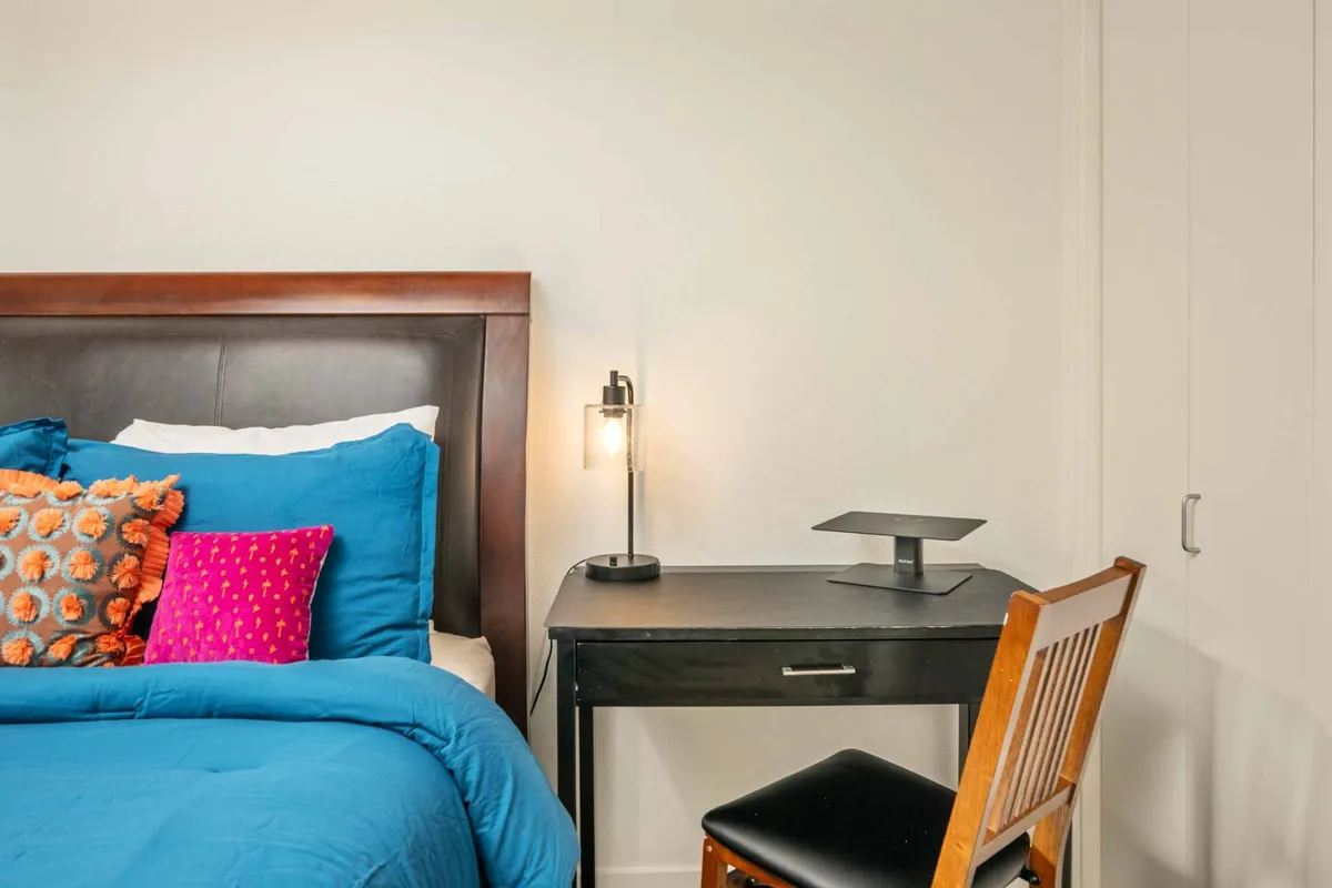 A cozy bedroom scene with a bed adorned with colorful pillows. Next to the bed is a dedicated desk complete with a lamp and comfortable chair, creating a productive and inviting workspace within a private space. Natural light streams in, enhancing the room’s warmth and making it ideal for both rest and focused work.
