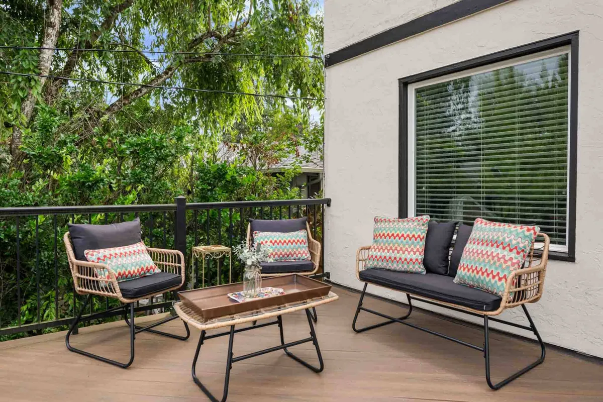 A sunny, comfortable exterior seating area with rattan chairs adorned with colorful cushions, a wooden table, and abundant greenery in the background. The space feels casual and inviting, suitable for both socializing and quiet relaxation.